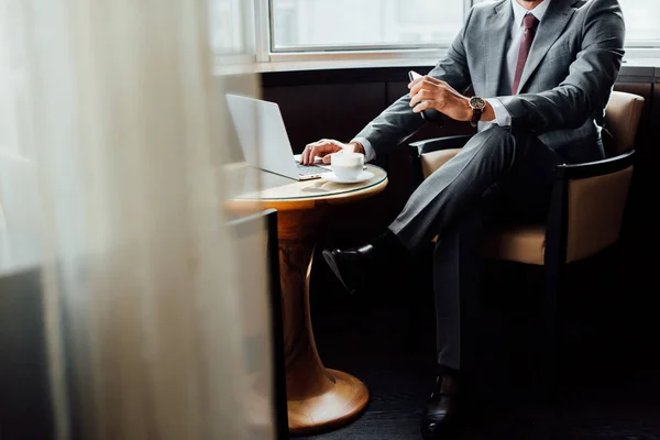 Vista recortada del hombre de negocios sosteniendo el teléfono inteligente mientras usa el ordenador portátil - foto de stock