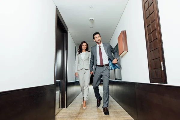 Confident businessman and happy businesswoman in formal wear holding hands and walking in hotel corridor — Stock Photo