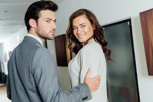 Hombre de negocios confiado tocando la mano de una alegre mujer de negocios en ropa formal mientras está de pie en el pasillo del hotel - foto de stock