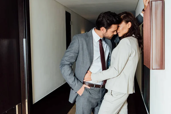 Homem bonito em terno de pé com a mão no bolso e beijando mulher atraente no corredor do hotel — Fotografia de Stock