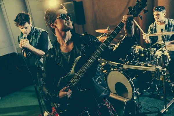 Enfoque selectivo de guapo guitarrista en gafas de sol tocando guitarra eléctrica cerca de banda de rock - foto de stock