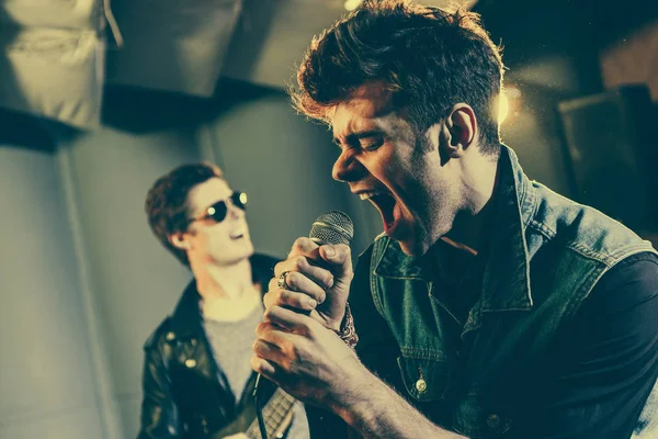 Selective focus of stylish singer singing in microphone near rock band — Stock Photo