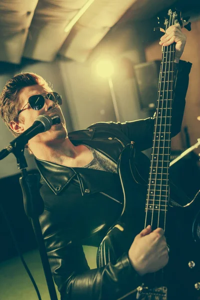 Beau guitariste en lunettes de soleil chantant chanson au microphone tout en jouant de la guitare électrique — Photo de stock