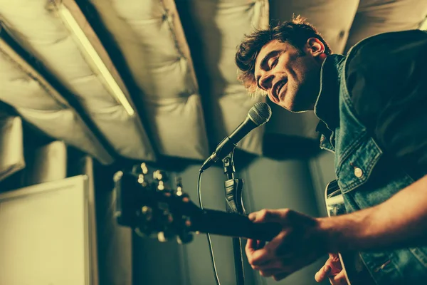 Foco seletivo do cantor alegre cantando em microfone enquanto toca guitarra acústica — Fotografia de Stock