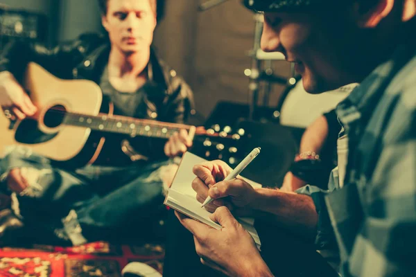 Foco seletivo de compositor alegre escrevendo em notebook perto de amigo tocando guitarra acústica — Fotografia de Stock