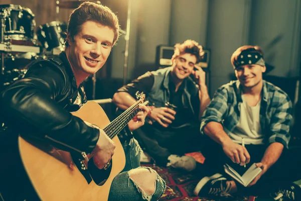 Enfoque selectivo de músico tocando la guitarra acústica y mirando a la cámara cerca de amigos felices sentados con las piernas cruzadas — Stock Photo