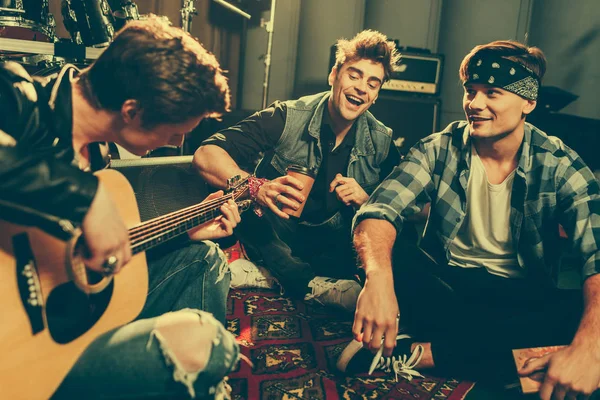 Selective focus of smiling friends sitting with crossed legs near musician playing acoustic guitar — Stock Photo