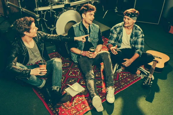 Overhead view of cheerful rock band holding paper cups and talking while sitting on carpet — Stock Photo