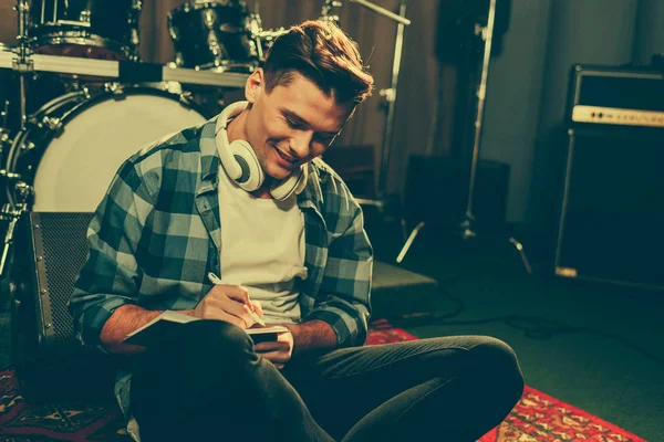 Cheerful composer in headphones writing in notebook while sitting near drums — Stock Photo