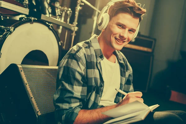 Alegre joven compositor escuchando música en auriculares mientras escribe en un cuaderno - foto de stock