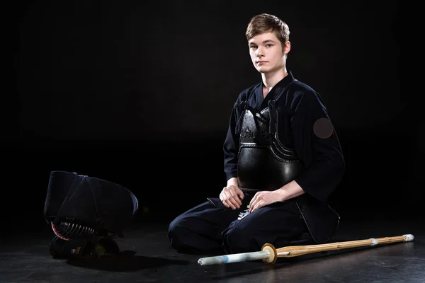 Handsome young man in kendo armor with bamboo sword sitting on black — Stock Photo