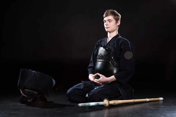 Young man in kendo armor sitting with closed eyes on black — Stock Photo