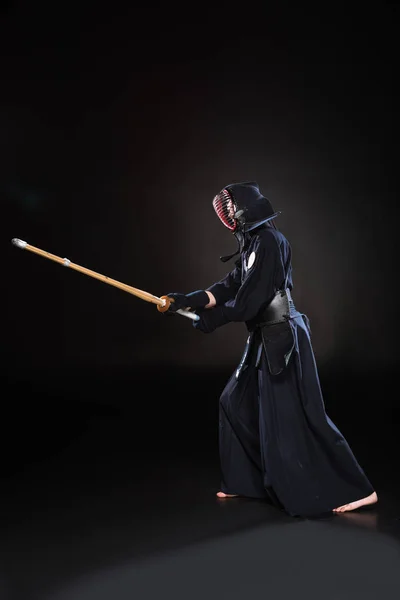 Vista lateral de kendo fighter en armadura practicando con espada de bambú sobre negro - foto de stock