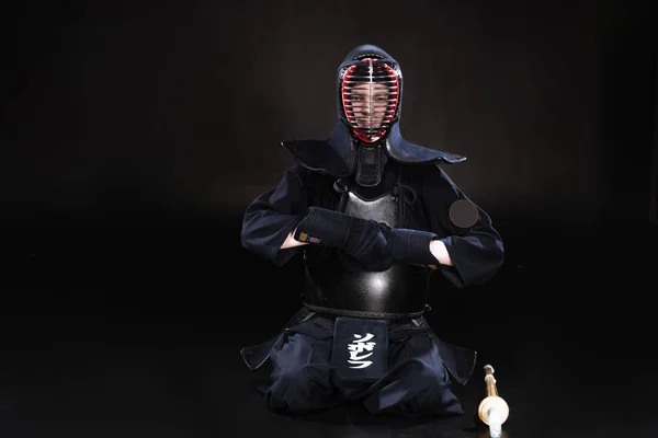 Kendo fighter in helmet sitting on floor and taking off gloves on black — Stock Photo