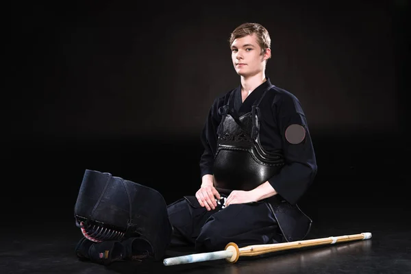 Young man in kendo armor with helmet and bamboo sword sitting on black — Stock Photo