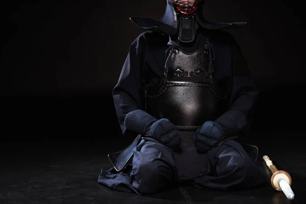 Vue partielle du combattant kendo en casque avec épée en bambou assis sur noir — Photo de stock