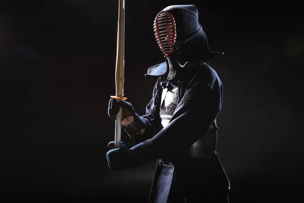 Kendo fighter in helmet holding bamboo sword on black — Stock Photo