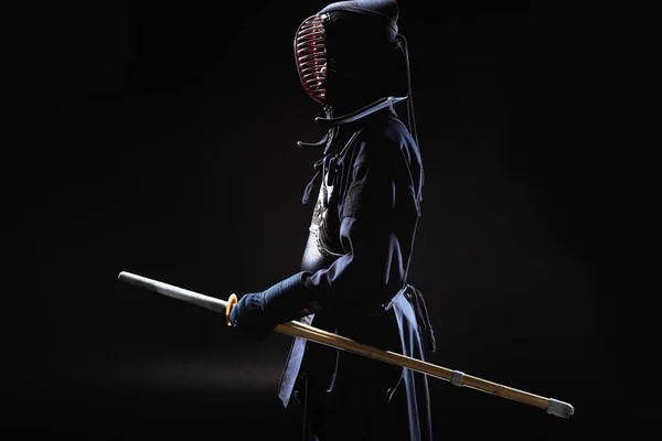 Vue latérale du combattant kendo en casque traditionnel tenant épée de bambou sur noir — Photo de stock