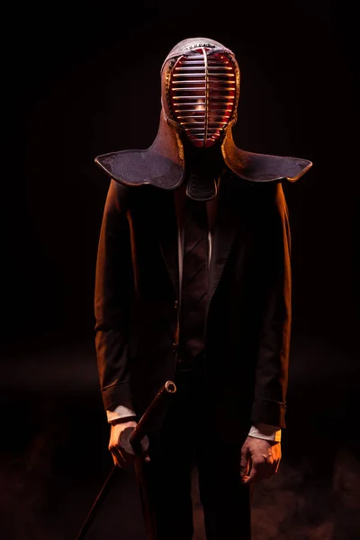 Hombre de traje y casco kendo sosteniendo espada de bambo en negro - foto de stock