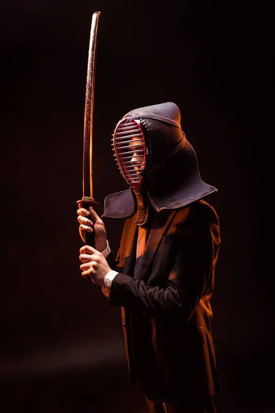 Kendo lutador em desgaste formal e capacete segurando espada de bambu no escuro — Fotografia de Stock