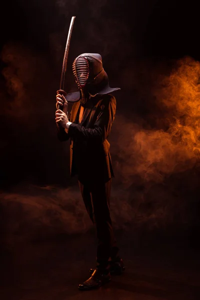 Visão de comprimento total do lutador kendo em desgaste formal e capacete segurando espada de bambu no escuro — Fotografia de Stock