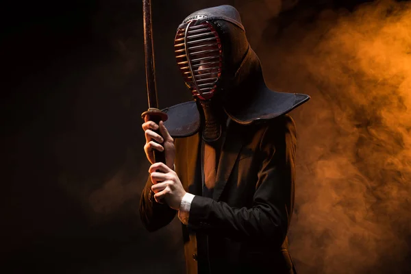 Combatiente Kendo en desgaste formal y casco sosteniendo espada de bambú en la oscuridad - foto de stock