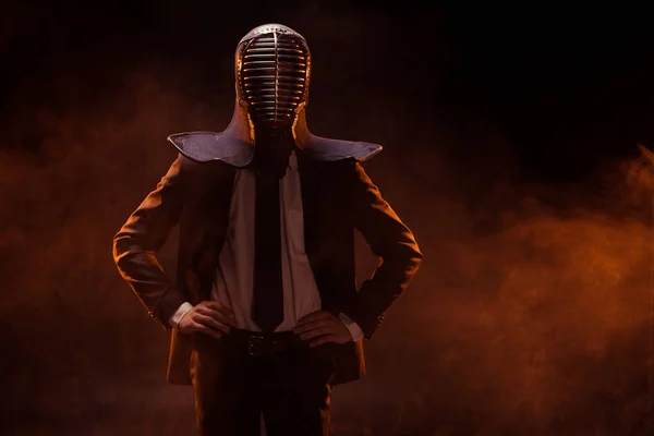 Kendo luchador en desgaste formal y casco de pie con los brazos akimbo en la oscuridad - foto de stock