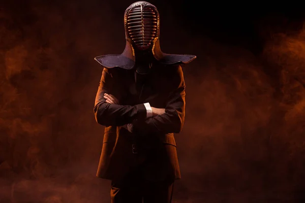 Combattant Kendo en tenue formelle et casque debout avec les bras croisés sur sombre — Photo de stock