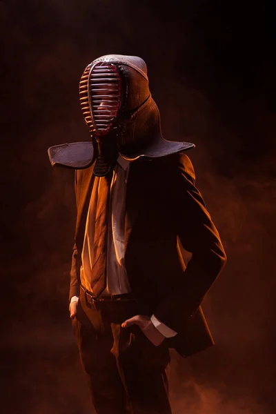 Confident kendo fighter in formal wear and helmet standing with hands in pockets on dark — Stock Photo