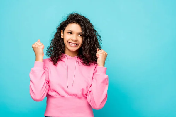 Happy curly african american girl smiling while celebrating triumph isolated on blue — Stock Photo