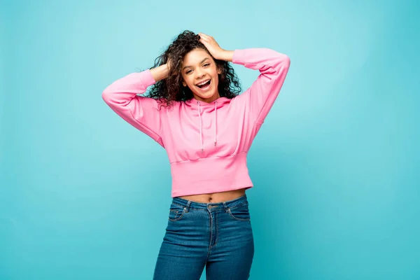 Cheerful curly african american woman touching hair isolated on blue — Stock Photo