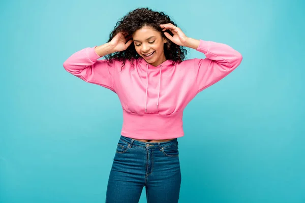 Happy curly african american woman touching hair isolated on blue — Stock Photo