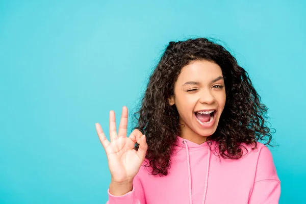 Fröhliche lockige afrikanisch-amerikanische Mädchen zwinkernden Augen, während zeigt ok Zeichen isoliert auf blau — Stockfoto