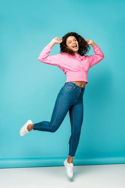 Happy curly african american girl in jeans jumping on blue — Stock Photo