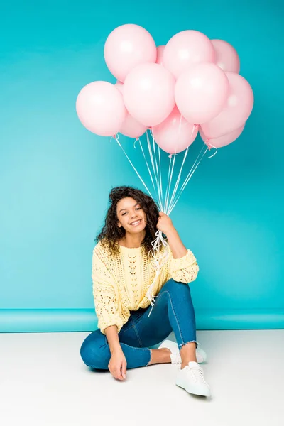 Pretty curly african american girl sitting with pink air balloons on blue — Stock Photo