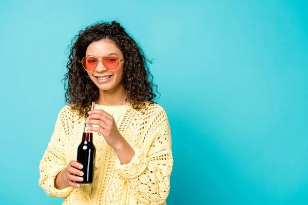 Cheerful african american girl in sunglasses holding bottle with straw on blue — Stock Photo