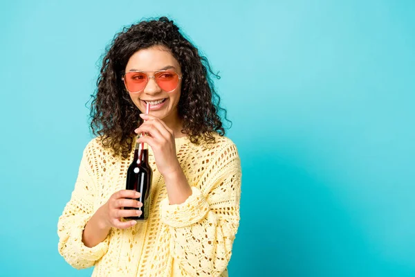 Fröhliche lockige afrikanisch-amerikanische Mädchen mit Sonnenbrille hält Flasche mit Stroh auf blau — Stockfoto