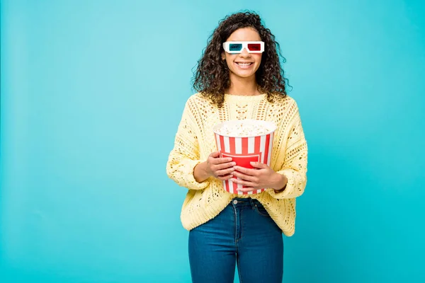 Joyeuse femme afro-américaine bouclée tenant seau de maïs soufflé et souriant dans des lunettes 3D sur bleu — Photo de stock