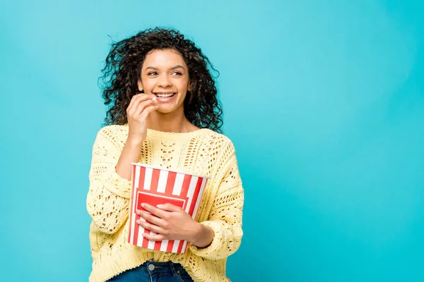 Fröhliche lockige Afroamerikanerin isst Popcorn isoliert auf blau — Stockfoto