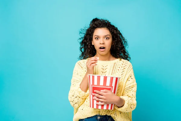 Schockiertes afrikanisches Mädchen isst Popcorn isoliert auf blauem Grund — Stockfoto