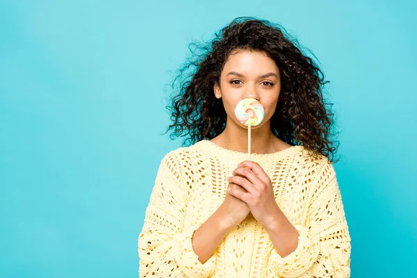 Curly african american girl covering mouth with lollipop isolated on blue — Stock Photo