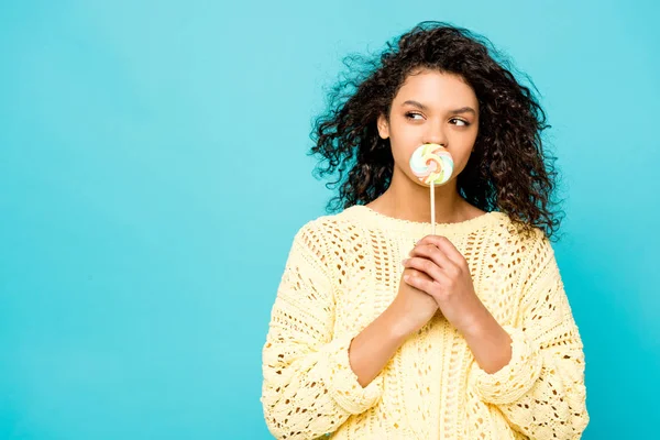 Dreamy curly african american girl covering mouth with lollipop isolated on blue — Stock Photo