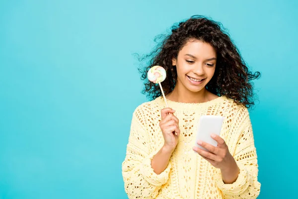Happy curly african american girl holding lollipop and using smartphone isolated on blue — Stock Photo