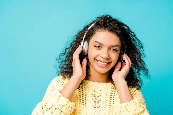 Feliz encaracolado menina americana africana ouvindo música em fones de ouvido e olhando para a câmera isolada no azul — Fotografia de Stock