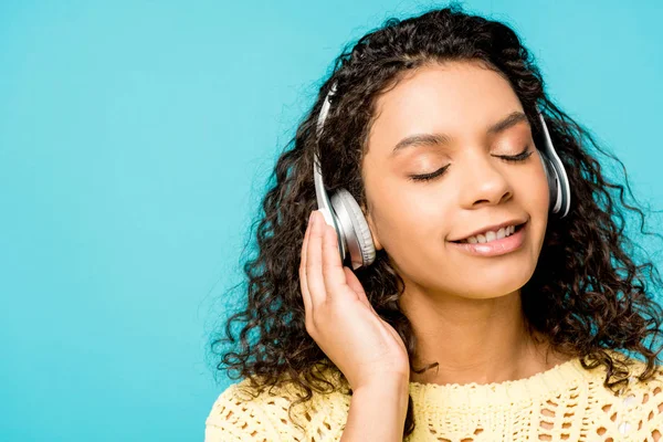 Alegre encaracolado afro-americano menina ouvindo música em fones de ouvido com olhos fechados isolados em azul — Fotografia de Stock