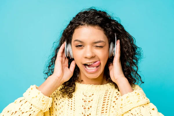 Atraente encaracolado menina americana africana ouvir música em fones de ouvido e mostrando língua isolada no azul — Fotografia de Stock