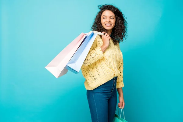 Счастливая кудрявая африканская девушка, держа в руках сумки с покупками, стоя на голубом фоне — стоковое фото