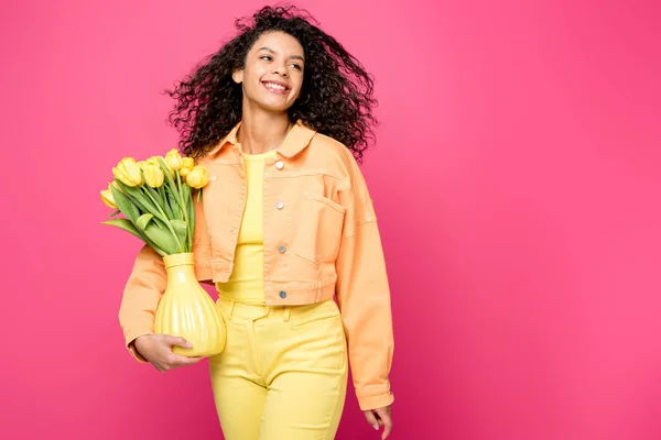 Glückliches afrikanisch-amerikanisches Mädchen hält Vase mit gelben Tulpen, während es isoliert auf purpurrotem Grund steht — Stockfoto