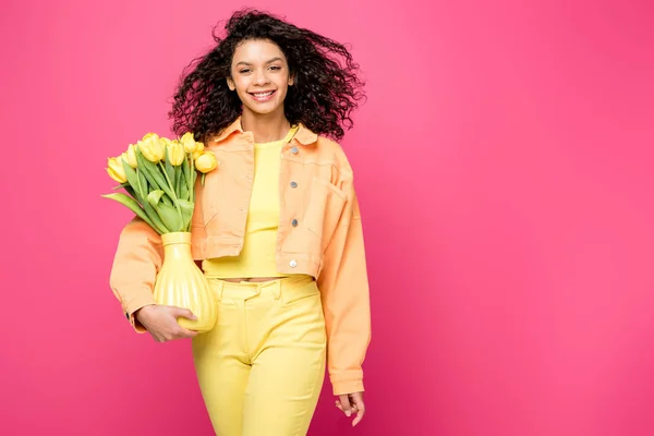 Sorridente menina afro-americana segurando vaso com tulipas amarelas, enquanto em pé isolado em carmesim — Fotografia de Stock