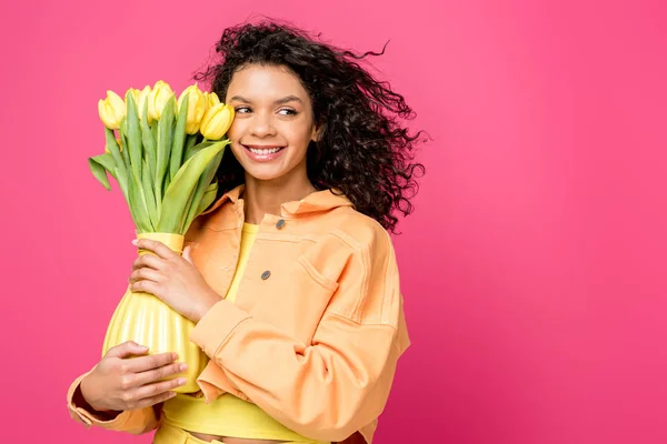 Attrayant sourire afro-américaine fille tenant vase avec des tulipes jaunes tout en se tenant isolé sur cramoisi — Photo de stock